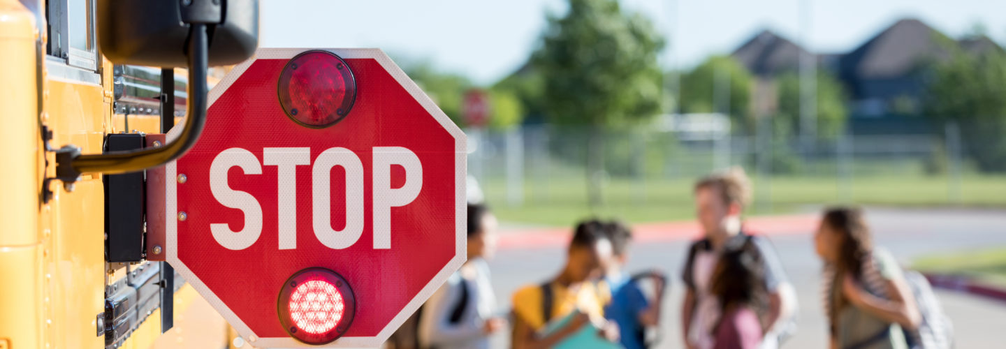A stop sign is attached to a bus, it is the focus, in the background, middle school students are standing near the bus
