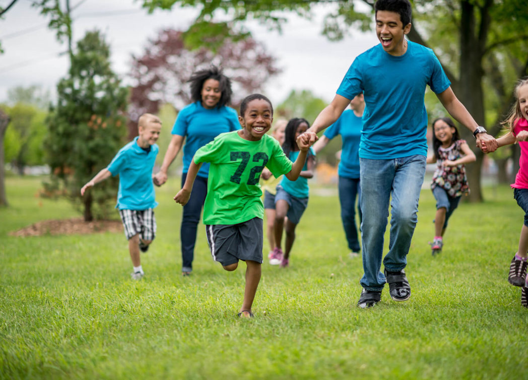 children in an afterschool program or summer camp are running at the camera, smiling and holding hands with the supervising adults who are also running