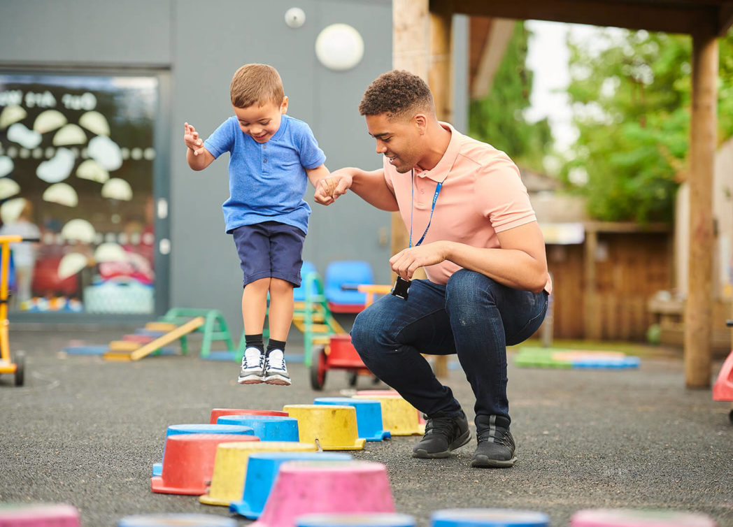 a pre school age child is mid jump at an early childhood care center while adult employee who is crouching holds the child's hand